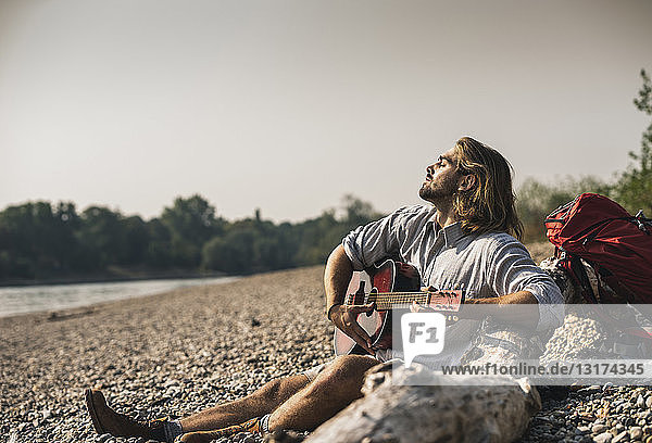 Young man sitting at the riverside playing guitar