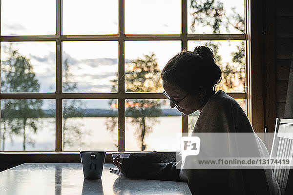 Finnland  Lappland  junge Frau sitzt am Fenster an einem See und schaut in ihr Tagebuch