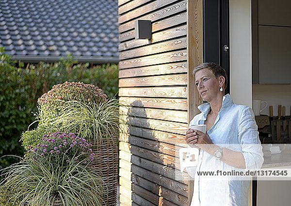 Nachdenkliche Frau mit Tasse Kaffee vor ihrem Haus stehend