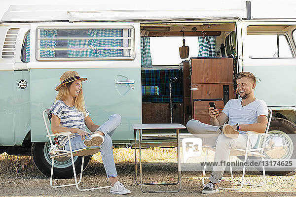 Happy young couple sitting on camping chairs at camper van with cell phone