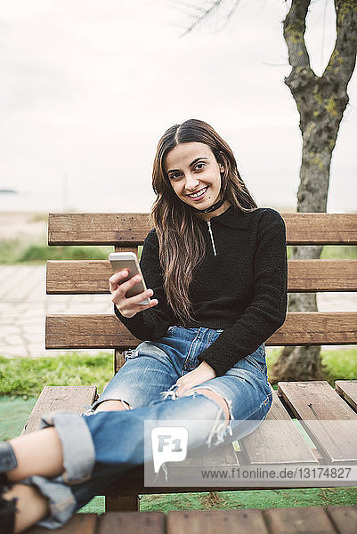 Porträt einer lächelnden jungen Frau mit Handy  die auf einer Bank im Freien sitzt