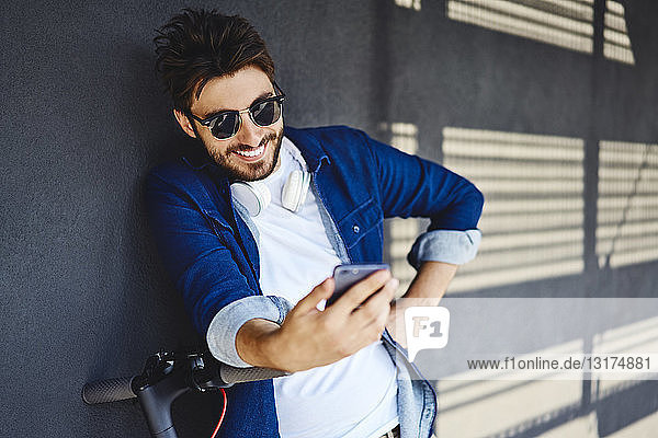 Porträt eines lächelnden jungen Mannes mit Elektroroller  der auf sein Handy schaut
