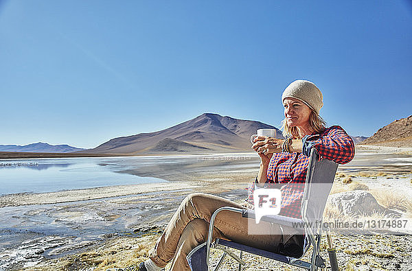 Bolivien  Laguna Colorada  Frau sitzt auf einem Campingstuhl am Seeufer und trinkt aus einer Tasse