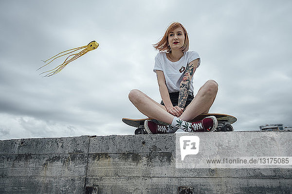 Junge Frau sitzt auf einer Betonwand auf einem Carver-Skateboard mit Drachen im Hintergrund