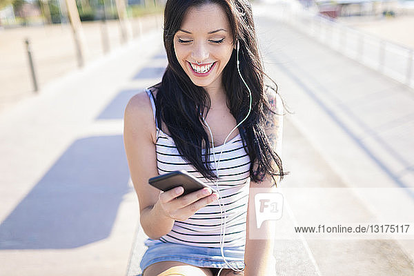 Glückliche junge Frau mit Nasenpiercing und Tätowierung mit Kopfhörer und Smartphone auf der Promenade