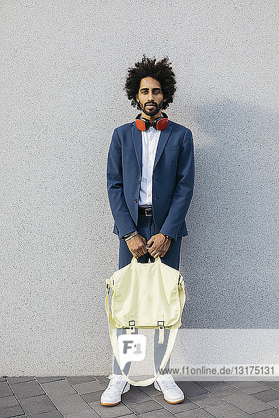 Portrait of stylish young businessman with bag and headphones at a wall