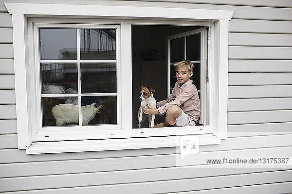 Porträt eines auf dem Fensterbrett sitzenden Jungen mit Jack Russel Terrier und Siamkatze  die aus dem offenen Fenster schauen