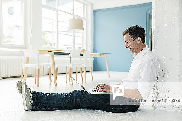 Smiling businessman sitting on the floor in office using laptop