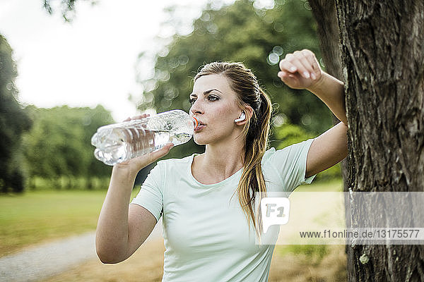 Sportliche junge Frau  die sich in einem Park an einen Baum lehnt und aus der Flasche trinkt