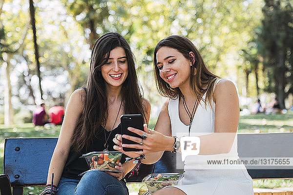 Girl friends sitting on a park bench looking at smartphone