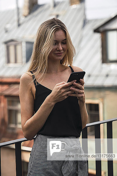 Porträt einer lächelnden blonden Frau  die auf dem Balkon steht und auf ihr Handy schaut