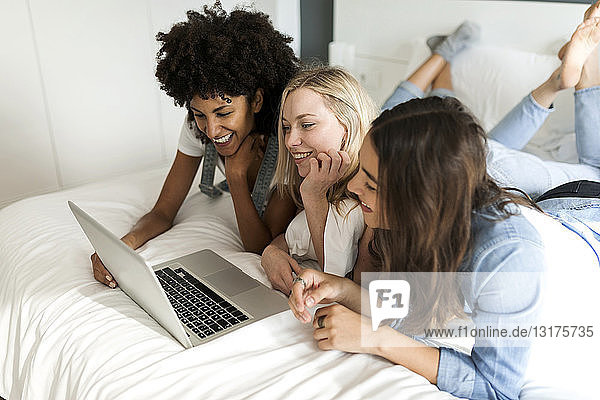 Three happy girlfriends lying on bed sharing laptop