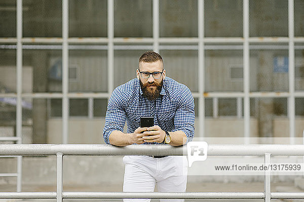 Portrait of bearded hipster businessman with smartphone leaning on railing