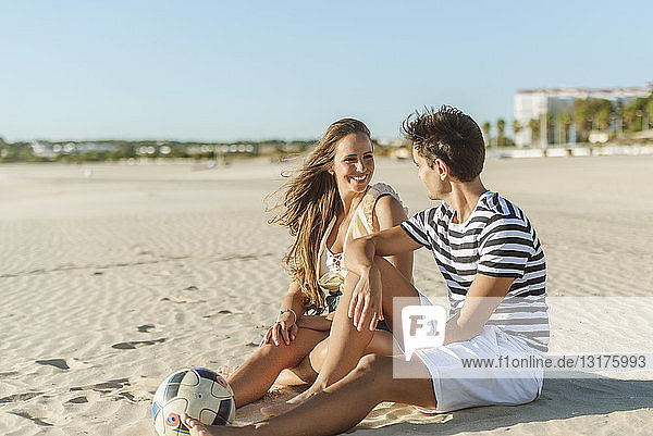 Lächelndes junges Paar sitzt mit einem Ball am Strand