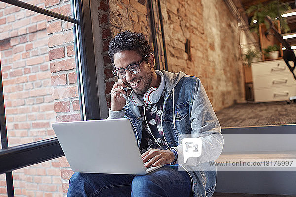 Junger Mann mit Kopfhörern und Laptop sitzt auf der Bürotreppe und telefoniert