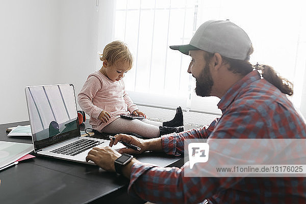 Vater arbeitet von zu Hause aus  benutzt einen Laptop  während seine Tochter auf dem Schreibtisch sitzt und spielt