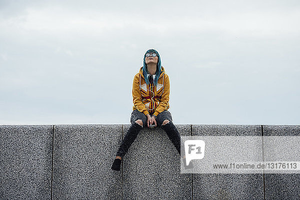 Young woman with dyed blue hair sitting on a wall looking up