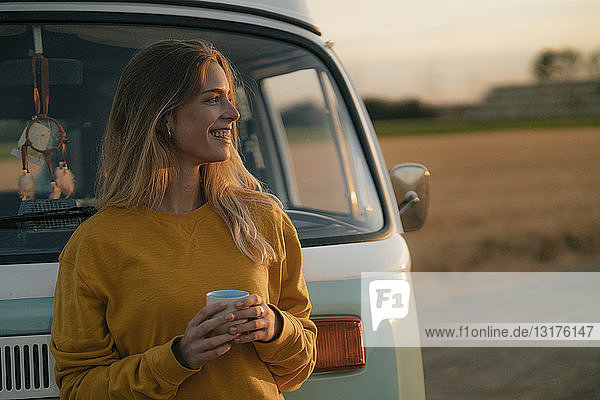 Glückliche junge Frau hält Becher am Wohnmobil in ländlicher Landschaft bei Sonnenuntergang