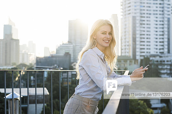 Blonde smiling business woman leaning onto handrail holding smartphone on city rooftop