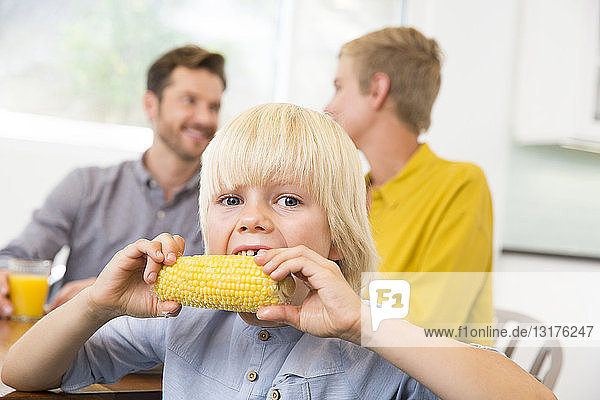 Junge isst Maiskolben in der Küche mit den Eltern im Hintergrund