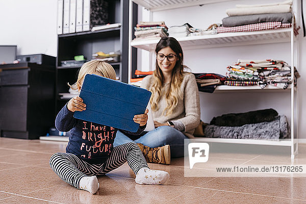 Mutter und kleine Tochter sitzen zu Hause mit Laptop und Tablett auf dem Boden