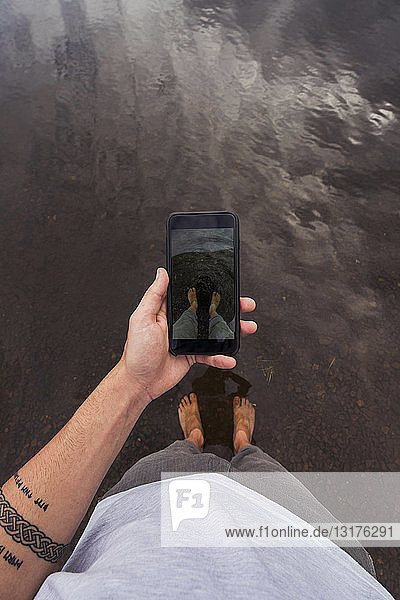Man taking a cell phone picture with feet in water of a lake