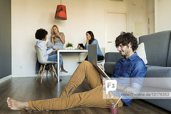 Smiling man sitting on floor using laptop with friends in background
