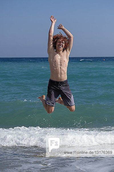 Porträt eines rothaarigen jungen Mannes,  der am Meeresufer in die Luft springt