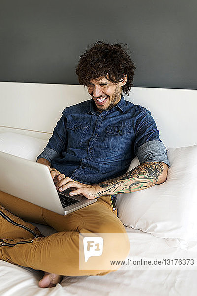 Glücklicher tätowierter Mann sitzt mit Laptop auf dem Bett