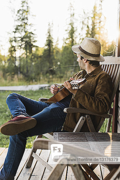 Young man sitting on veranda of a wood house  playing the ukulele
