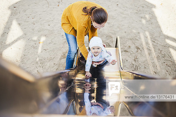 Lächelndes kleines Mädchen auf der Rutsche auf dem Spielplatz,  das von seiner Mutter gehalten wird