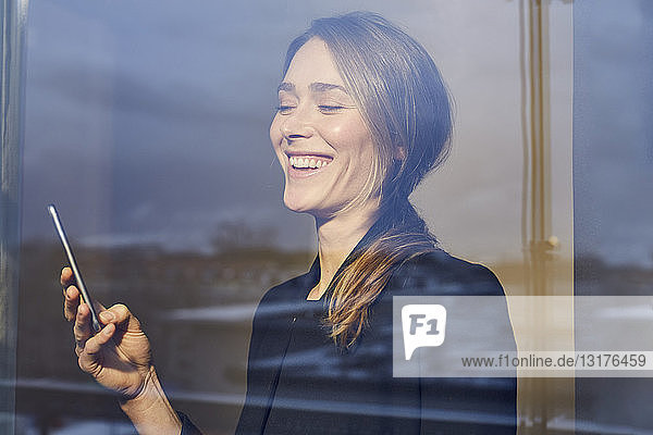 Porträt einer lachenden Geschäftsfrau hinter einer Fensterscheibe,  die auf ein Mobiltelefon schaut