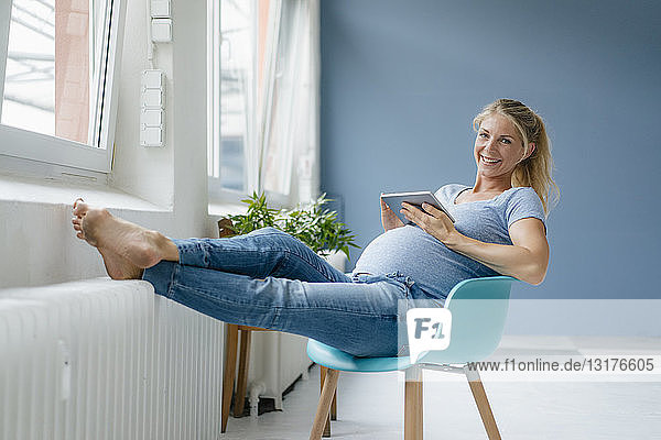 Porträt einer lächelnden schwangeren Frau  die auf einem Stuhl am Fenster sitzt und ein Tablett hält