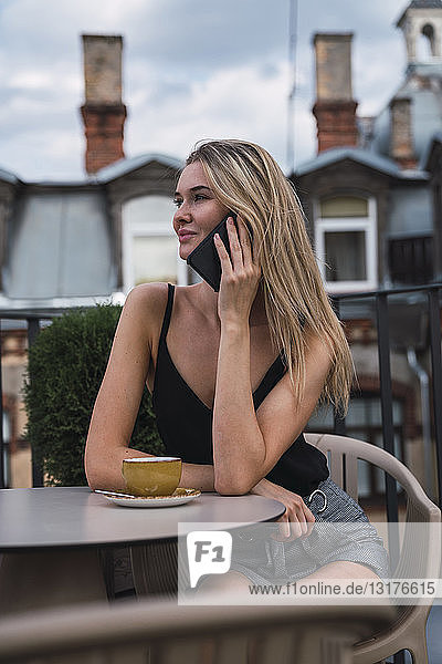 Lächelnde blonde Frau am Telefon  die auf der Dachterrasse bei einer Tasse Kaffee sitzt
