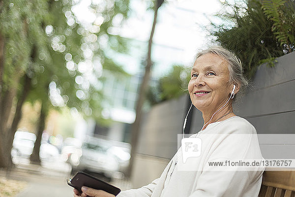 Lächelnde ältere Frau sitzt auf einer Bank im Freien mit Handy und Kopfhörern
