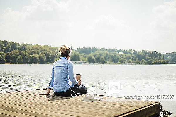 Woman sitting on jetty at a lake with headphones and takeaway coffee