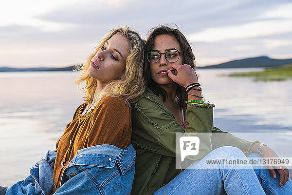 Finland  Lapland  two young women sitting back to back at the lakeside at twilight