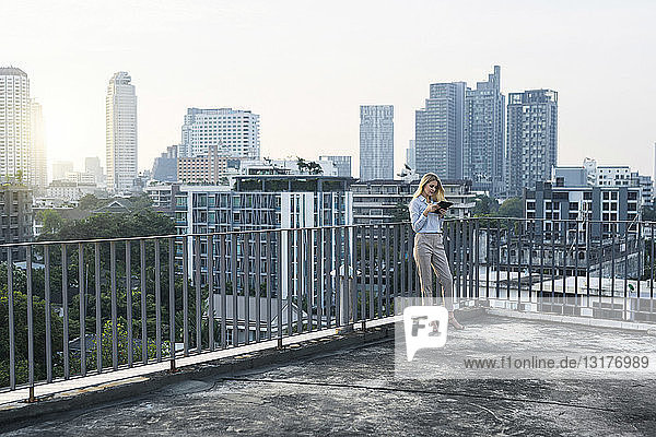 Blonde business woman leaning onto handrail holding tablet on city rooftop