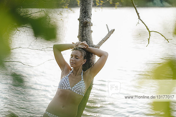 Relaxed woman wearing a bikini leaning against tree trunk at a lake