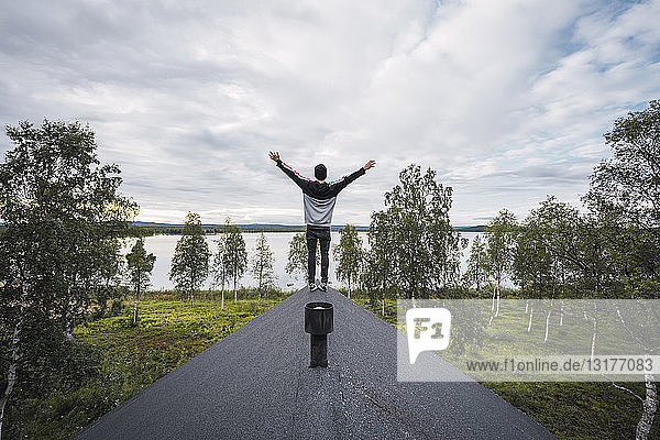 Finnland Lappland Mann steht mit erhobenen Armen auf dem Dach eines Hauses an einem See