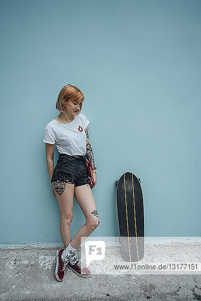 Coole junge Frau steht an türkisfarbener Wand neben Carver-Skateboard
