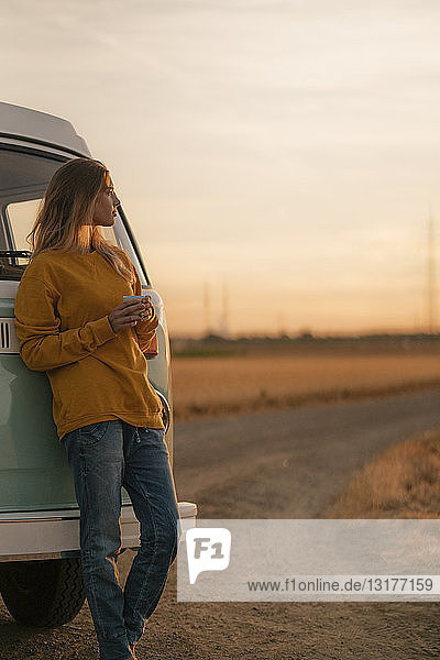 Junge Frau steht am Wohnmobil in ländlicher Landschaft bei Sonnenuntergang