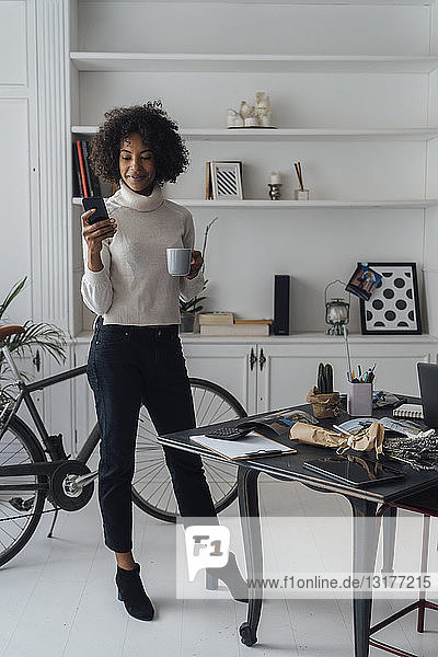 Mid adult freelancer standing in her home office  using smartphone  holding coffee cup