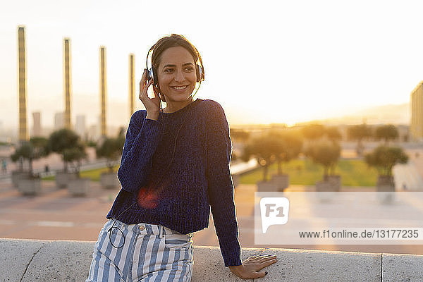 Spanien  Barcelona  Montjuic  lächelnde junge Frau  die bei Sonnenuntergang mit Kopfhörern Musik hört