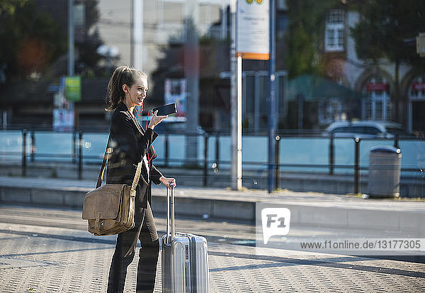Smiling young woman with luggage at tram station in the city using cell phone