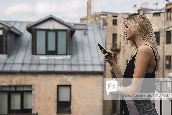 Blonde junge Frau steht auf Balkon und schaut auf Handy