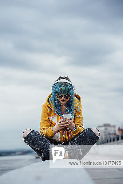Junge Frau mit blau gefärbten Haaren sitzt an einer Wand und hört mit Kopfhörern und Smartphone Musik