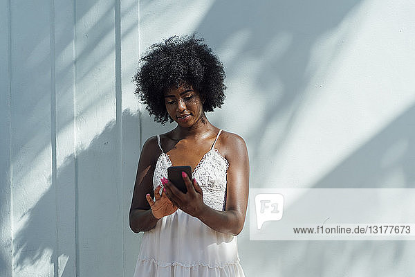 Junge Frau in weißem Kleid mit Mobiltelefon an einer Wand