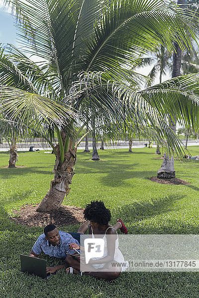 USA  Florida  Miami Beach  junges Paar mit Tablet und Laptop auf einem Rasen in einem Park