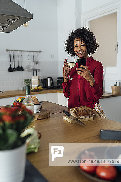 Frau trinkt Weißwein in ihrer Küche und benutzt ein Smartphone